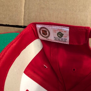 May include: Red baseball cap with a green brim. The inside of the cap has a white label with the NHL logo and text that reads "Official Licensed Product NHL", "The G.C.C. Quality Since 1903", "Cap", "80% Acrylic 20% Wool", "One Size Fits All", and "Made in Bangladesh".