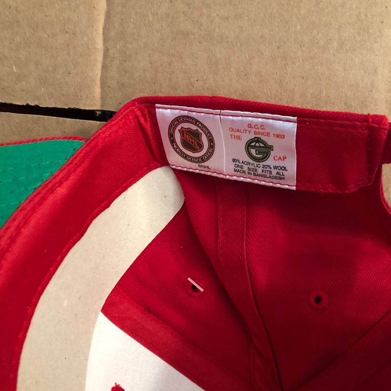 May include: Red baseball cap with a green brim. The inside of the cap has a white label with the NHL logo and text that reads "Official Licensed Product NHL", "The G.C.C. Quality Since 1903", "Cap", "80% Acrylic 20% Wool", "One Size Fits All", and "Made in Bangladesh".