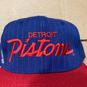 Op de afbeelding: Een blauwe en rode baseballpet met een wit pinstripe patroon. De pet heeft de woorden "DETROIT" en "Pistons" in rood geborduurd op de voorkant.