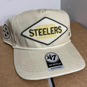 May include: A tan baseball cap featuring the Pittsburgh Steelers logo in black and gold. The patch displays the team name and "ESTABLISHED 1933". The hat has a curved peak and an adjustable strap.