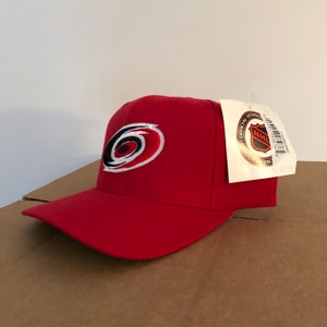 May include: Red baseball cap with a white and black Carolina Hurricanes logo. The cap has an adjustable strap in the back. The tag attached to the cap reads "Official Licensed Product NHL".