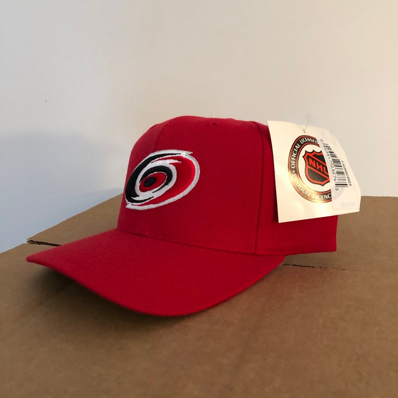 May include: Red baseball cap with a white and black Carolina Hurricanes logo. The cap has an adjustable strap in the back. The tag attached to the cap reads "Official Licensed Product NHL".