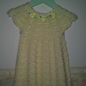 May include: A crocheted baby dress in a pale yellow color with a scalloped hem. The dress has a row of yellow fabric flowers around the neckline and a few more flowers around the hem.