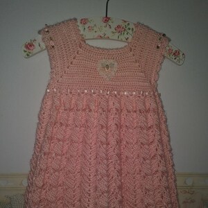 May include: A pink crocheted baby dress with a scalloped hem and floral embellishments. The dress has a delicate lace pattern and a small heart-shaped applique on the bodice.