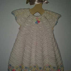 May include: A white crocheted baby dress with a scalloped hem and colorful ribbon flowers. The dress has short sleeves and a round neckline.