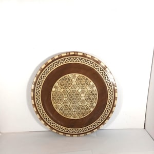 Puede incluir: Un plato redondo decorativo de madera con un centro marrón oscuro y un borde marrón más claro. El centro presenta un intrincado patrón geométrico en color crema. El borde exterior tiene un diseño geométrico repetitivo en crema y marrón.