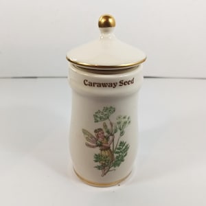 Può includere: Un barattolo di spezie in ceramica color crema con coperchio e dettagli dorati. Il barattolo è etichettato "Caraway Seed" in testo marrone e presenta un'illustrazione di una fata. Il coperchio ha un pomello dorato.