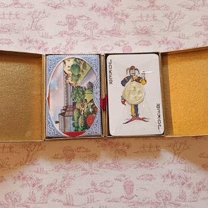 May include: An open gold-colored box containing two decks of playing cards. One deck features a colorful illustration, while the other displays a joker card with a gold coin. The box sits on a pink floral patterned surface.