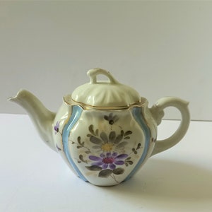 May include: A white porcelain teapot with a floral design in shades of purple, blue, and green. The teapot has a gold trim around the lid and a blue and white striped design on the body.