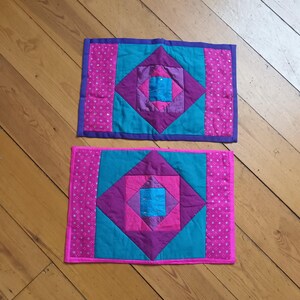 May include: Two colorful quilted placemats. Each features a central diamond pattern in teal, purple, and pink, with a small square of blue in the center. The borders are pink with a dotted pattern and purple trim.