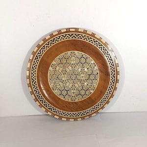 Puede incluir: Plato redondo de madera con intrincados diseños geométricos. El plato presenta un diseño central de formas entrelazadas, rodeado por un borde de segmentos de madera claros y oscuros alternados. El borde exterior tiene un patrón triangular repetitivo.