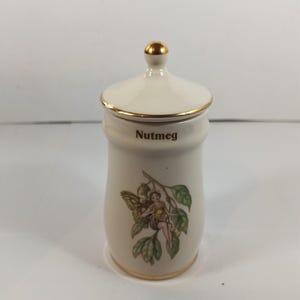 Può includere: Barattolo per spezie di noce moscata in ceramica color crema con coperchio e finiture dorate. Il barattolo presenta un'illustrazione di fata e la scritta "Nutmeg" in lettere marroni. Il coperchio ha un pomello dorato.
