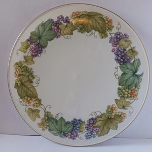 Könnte beinhalten: Ein weißer Keramikteller mit goldfarbenem Rand zeigt ein kreisförmiges Design aus grünen Weinreben mit violetten, grünen und gelben Trauben.