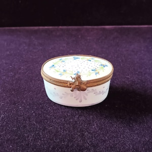 May include: An oval, white porcelain trinket box with a floral design and gold-colored metal trim. The lid features blue flowers and a dotted pattern. A small, decorative clasp secures the lid.