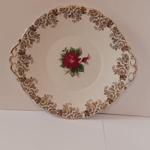 May include: A white ceramic serving plate with a gold rim and floral design. The center of the plate features a red rose with green leaves.