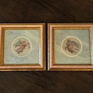 Könnte beinhalten: Zwei gerahmte Kunstwerke mit Engelsillustrationen. Jedes Kunstwerk hat einen hellblauen Hintergrund mit einem kreisförmigen Engelbild. Die Rahmen sind gold- und rosafarben und bieten eine klassische Ästhetik. Ideal für die Inneneinrichtung.
