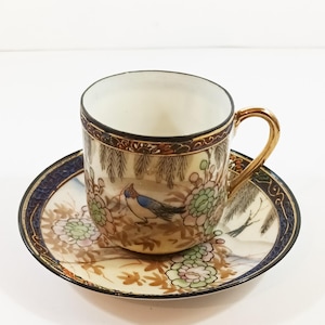 Op de afbeelding: Een vintage kop en schotel set. De kop en schotel hebben een gedetailleerd bloemen- en vogelontwerp in bruin-, blauw- en groentinten. De kop heeft een gouden handvat en rand, en de schotel heeft een donkerblauwe rand.