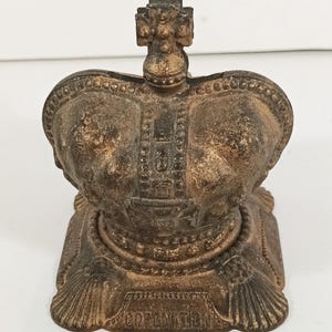 Op de afbeelding: Een bronskleurig kroonvormig object met het woord "CORONATION" aan de basis. De kroon heeft een gedetailleerd ontwerp met een kruis erop. Het object is waarschijnlijk een decoratief stuk of souvenir.