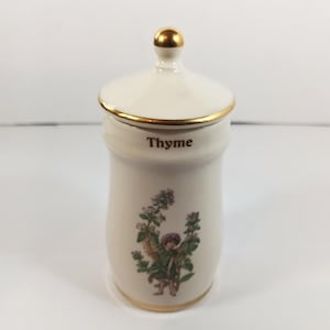 Może przedstawiać: Kremowy ceramiczny słoik na przyprawy z pokrywką i złotym wykończeniem. Na przodzie nadrukowano napis "Thyme". Mała ilustracja wróżki z gałązkami tymianku znajduje się z przodu. Pokrywka ma złoty uchwyt.