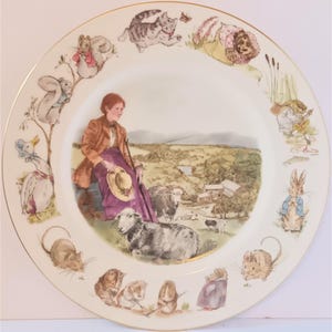 Może przedstawiać: Biały ceramiczny talerz ze złotą obwódką przedstawia kolorową ilustrację kobiety siedzącej na wzgórzu z owcami. Talerz jest ozdobiony obramowaniem z postaciami zwierząt z książek dla dzieci Beatrix Potter.