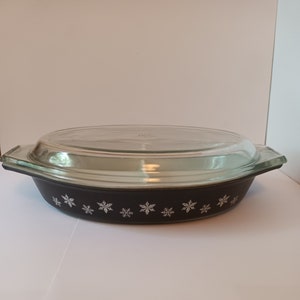 Puede incluir: Una fuente para hornear ovalada negra con tapa de vidrio. La fuente tiene un patrón de copo de nieve blanco alrededor del borde.