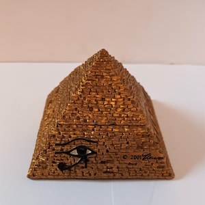 May include: A gold-coloured pyramid-shaped trinket box with a black eye of Horus design on the front. The box is inscribed with the copyright symbol and the year 2001.