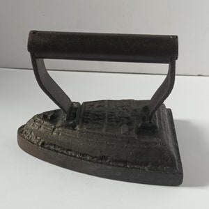 Puede incluir: Una plancha de hierro fundido vintage con acabado negro y mango. La plancha tiene una base redondeada y un diseño en relieve en la parte superior.