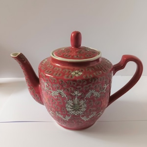 Peut inclure: Une théière en céramique rouge avec un motif floral. La théière a un couvercle et un bec verseur. Le motif floral est en blanc et or.
