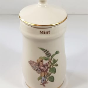 May include: Cream ceramic mint jar with gold trim and lid. The lid has a gold knob. The word "Mint" is printed on the jar. A fairy illustration is on the front, holding a sprig of mint.