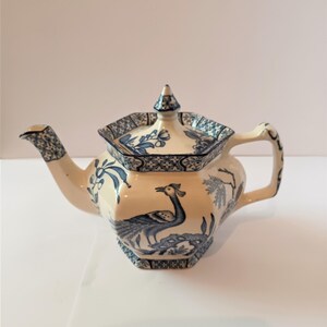 May include: Cream-colored ceramic teapot with blue floral and peacock designs. The teapot has a hexagonal shape, a curved spout, and a handle with a decorative pattern. The lid is topped with a small finial. A classic piece for tea lovers.