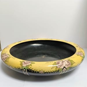 Puede incluir: Un cuenco decorativo ovalado con interior negro y borde amarillo. El borde está adornado con rosas rosas y hojas verdes. El exterior del cuenco es negro, creando un contraste llamativo con el amarillo y el diseño floral.