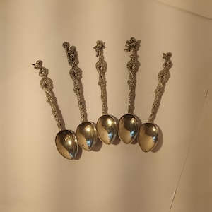 May include: Five decorative silver spoons with detailed handles and polished bowls are displayed on a white surface. Each spoon has a different decorative figure on the handle, including cherubs and floral designs. The spoons are likely for display or collecting.