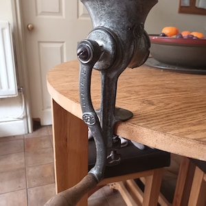 May include: A vintage cast iron manual food grinder with a wooden handle. The grinder is silver with black accents and has a crank handle. The grinder is sitting on a wooden table.