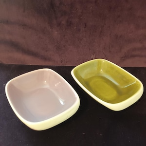 Può includere: Due piccole ciotole rettangolari in ceramica con angoli arrotondati. Una presenta un interno liscio grigio chiaro, mentre l'altra ha un interno verde oliva lucido. Entrambe le ciotole hanno una finitura esterna color crema.