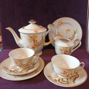 May include: A white porcelain tea set with a dragon design. The set includes a teapot, creamer, sugar bowl, two teacups, and two saucers. The dragon is depicted in gold, orange, and brown colors.