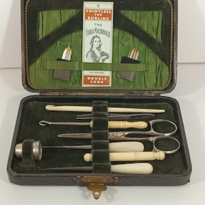 May include: An open antique sewing kit with various tools, including scissors, needles, and a thimble. The kit is housed in a dark leather case with a green velvet interior. The kit includes a card that reads "Pointless for Burling" and "The Flora Macdonald".
