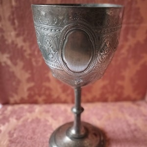 Könnte beinhalten: Ein silberner Kelch mit einem verzierten Design. Der Kelch hat eine runde Schale mit einer erhöhten Mitte und einen Stiel mit einer runden Basis.