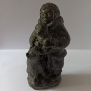 May include: A dark grey sculpture of a person holding a child and a dog. The person wears a hooded coat with a fur trim. The sculpture is detailed and appears to be made of stone or a similar material.