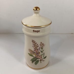 Puede incluir: Tarro de especias de cerámica color crema con tapa y ribete dorado. El tarro lleva la inscripción "Sage" en texto marrón y presenta una ilustración floral con un hada. La tapa tiene un pomo dorado.