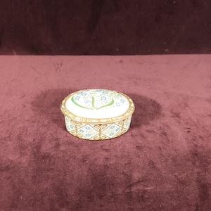 Puede incluir: Una caja de baratijas ovalada de porcelana con tapa. La caja presenta un borde dorado y está decorada con motivos florales azules y detalles verdes. El fondo es de color burdeos oscuro.
