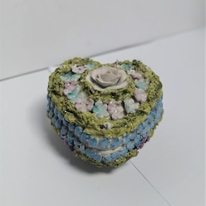 May include: Heart-shaped trinket box with a floral design. The box has a light green top with a central rose and small pink, blue, and white flowers. The sides are blue with a textured appearance, creating a decorative piece.