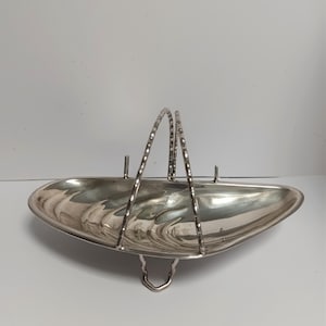 May include: A silver-colored, oval-shaped serving dish with a handle. The dish has a reflective surface and a decorative handle. The handle is made of two arched pieces with a textured design. The dish has a small, curved foot.