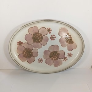 Puede incluir: Plato ovalado de cerámica con diseño floral. El plato presenta un fondo claro con flores rosas y centros marrones. El borde es de un tono marrón más oscuro. Una pieza decorativa para servir alimentos.