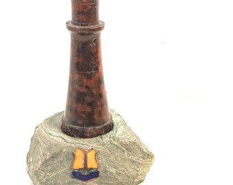 Puede incluir: Una figura de faro de color marrón moteado con una parte superior redondeada, colocada sobre una base de piedra natural gris y rugosa. La piedra presenta un pequeño emblema en forma de escudo con colores naranja y azul y el texto LANDS END.