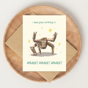 May include: A cream-colored greeting card featuring a brown, multi-legged creature illustration with yellow stars. The text reads: I hope your birthday is AMAZE! AMAZE! AMAZE! The card is placed on a brown envelope and a round wooden tray.