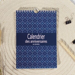 Op de afbeelding: Een blauw patroon verjaardagskalender met de tekst "Calendrier des anniversaires" ligt op een geweven placemat. Een potlood en drie paperclips zijn ook zichtbaar.