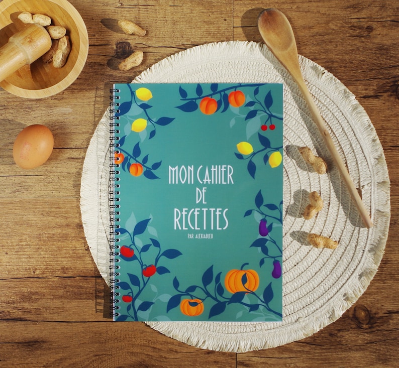 K&ouml;nnte beinhalten: Ein t&uuml;rkisfarbenes Notizbuch mit Spiralbindung, einem farbenfrohen Blumendesign und dem Text "Mon Cahier de Recettes par Alezandra".