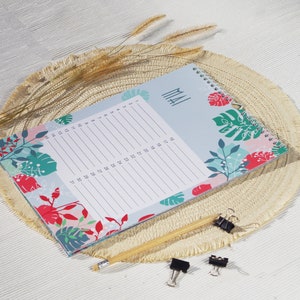 May include: A spiral-bound monthly planner with a light blue cover featuring red, green, and teal tropical leaf patterns. The page displays the text MAI and a numbered list from 1 to 31 for daily entries. A yellow pencil and two black binder clips are included.