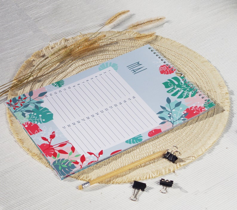 May include: A spiral-bound monthly planner with a light blue cover featuring red, green, and teal tropical leaf patterns. The page displays the text MAI and a numbered list from 1 to 31 for daily entries. A yellow pencil and two black binder clips are included.