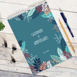 May include: A spiral-bound birthday calendar with a teal cover featuring a botanical pattern of leaves and branches in shades of dark blue, light blue, white, and peach. The text reads "CALENDRIER DES ANNIVERSAIRES" and "PAR ALEXIABLEU".
